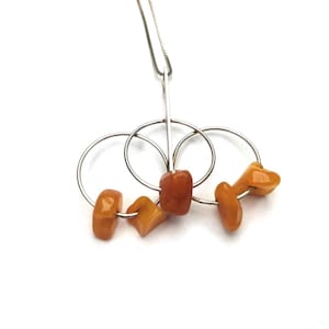 Peut inclure: Un collier en argent avec trois anneaux entrelacés, chacun orné d'une perle d'ambre. Le collier a une chaîne fine et une barre centrale. Les perles d'ambre sont de différentes nuances d'orange et de brun.