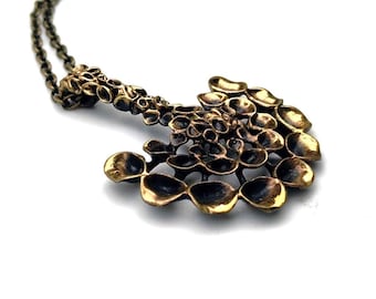 May include: A bronze-toned pendant necklace with a unique, organic design. The pendant features a series of interconnected, textured shapes, creating a flowing, abstract form. The chain is a matching bronze color, and the necklace is set against a white background.
