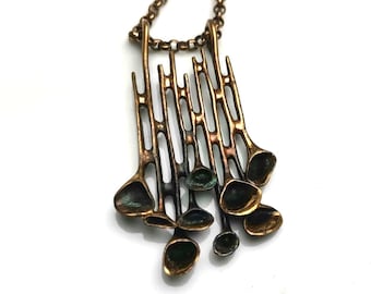 May include: A bronze-toned metal pendant necklace featuring a series of vertical, rod-like structures that terminate in small, cup-shaped forms. The metal has a darkened, oxidized finish with hints of green patina, suspended from a matching chain.
