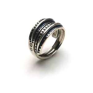 May include: A silver-toned multi-band ring featuring three distinct, stacked-style layers. The outer bands are decorated with small, dark, recessed circular and linear patterns, while the inner band remains smooth and polished in appearance.