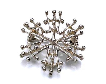 Könnte beinhalten: Eine silberne Brosche in Form einer Schneeflocke mit einem filigranen, komplizierten Design. Die Brosche besteht aus vielen kleinen, runden Perlen, die durch dünne Drähte verbunden sind.