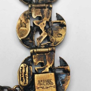 May include: A bronze bracelet with a geometric design. The bracelet is made up of several circular links that are connected by hinges. The links are decorated with a pattern of interlocking shapes. The bracelet is stamped with the words "Bronze Finland" and "Tommi Laine".