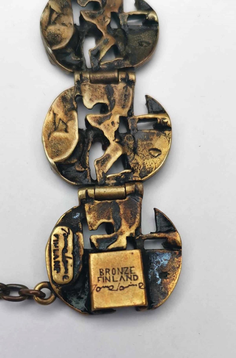 May include: A bronze bracelet with a geometric design. The bracelet is made up of several circular links that are connected by hinges. The links are decorated with a pattern of interlocking shapes. The bracelet is stamped with the words "Bronze Finland" and "Tommi Laine".