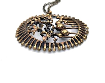 May include: A gold-toned pendant necklace with a circular design featuring a textured, abstract pattern. The pendant has a raised, organic shape in the center.