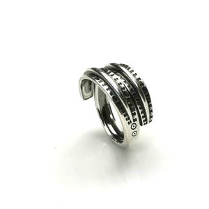 Könnte beinhalten: Ein silberfarbener Ring mit einem einzigartigen, geschichteten Design. Der Ring hat mehrere Bänder mit einem strukturierten Muster und kleinen, dunklen Akzenten. Das offene Design ermöglicht eine verstellbare Passform. Der Ring wird vor weißem Hintergrund gezeigt.