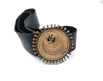 May include: A black leather belt with a large, ornate gold-colored buckle. The buckle is circular with intricate patterns and small, decorative studs around the edge. The belt is wide and appears to be made of textured leather.