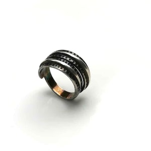 May include: A silver ring with a unique design. The ring features three stacked bands with a textured, ridged appearance. The bands are accented with small, dark details, creating a contrast against the silver. The ring is set against a white background.