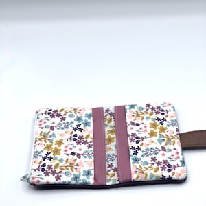 May include: A small floral print wallet with a zipper closure and a brown leather strap. The wallet has a pink and white floral pattern with green leaves.