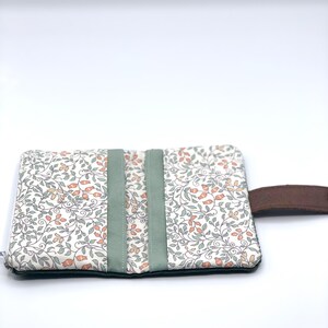 May include: A white fabric pouch with a floral pattern and green trim. The pouch has a brown leather strap.
