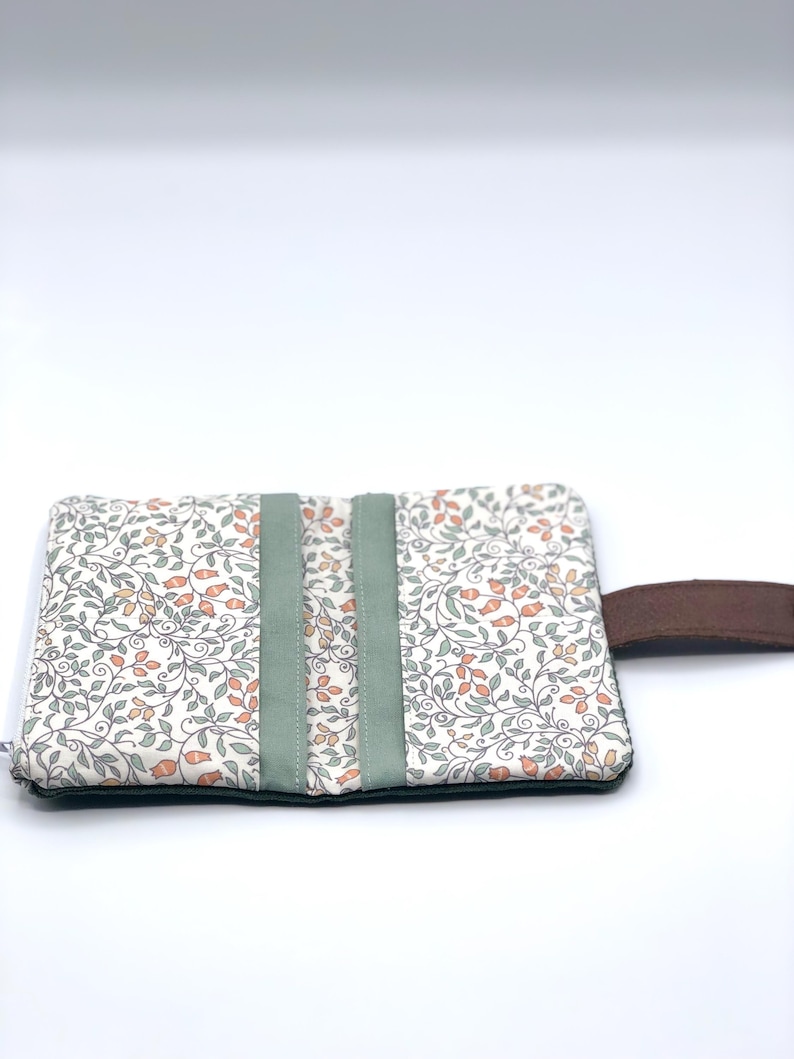 May include: A white fabric pouch with a floral pattern and green trim. The pouch has a brown leather strap.