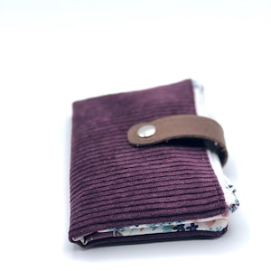 May include: A small, burgundy corduroy wallet with a brown leather flap and a silver snap closure. The wallet is folded closed and the inside lining is visible, featuring a floral pattern.