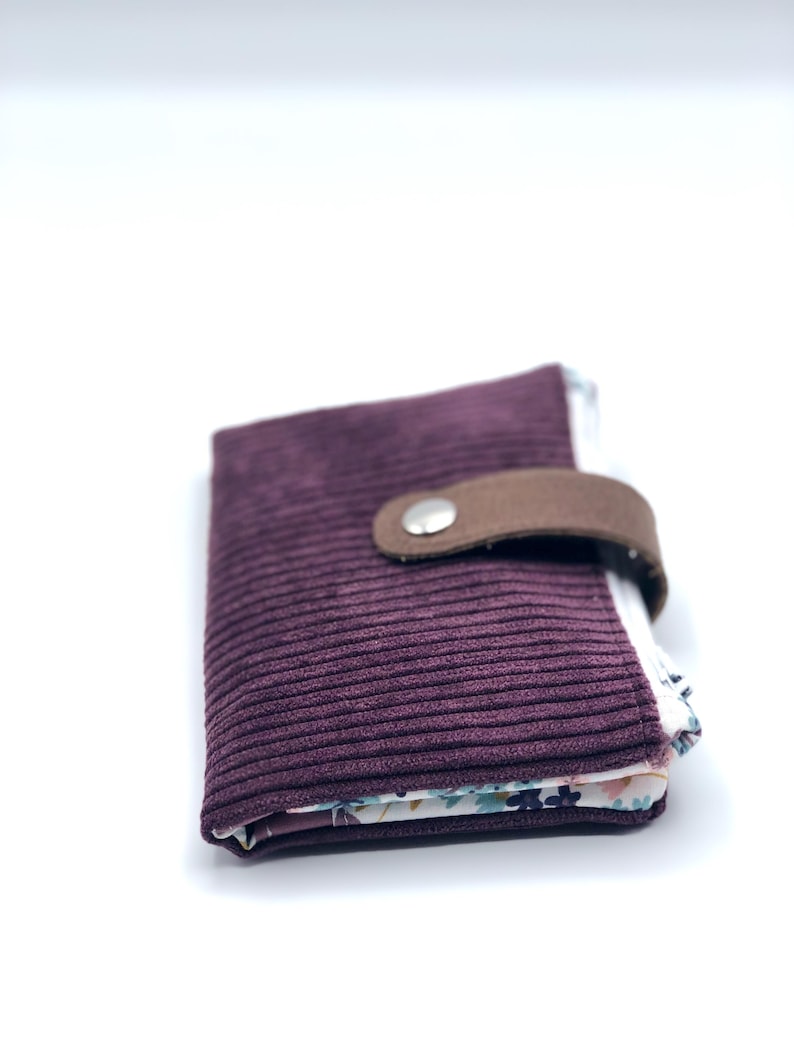 May include: A small, burgundy corduroy wallet with a brown leather flap and a silver snap closure. The wallet is folded closed and the inside lining is visible, featuring a floral pattern.