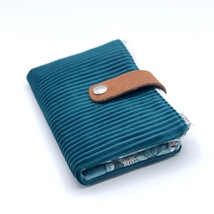 May include: A teal blue corduroy wallet with a brown leather strap and a silver button closure. The inside of the wallet is lined with a floral print fabric.