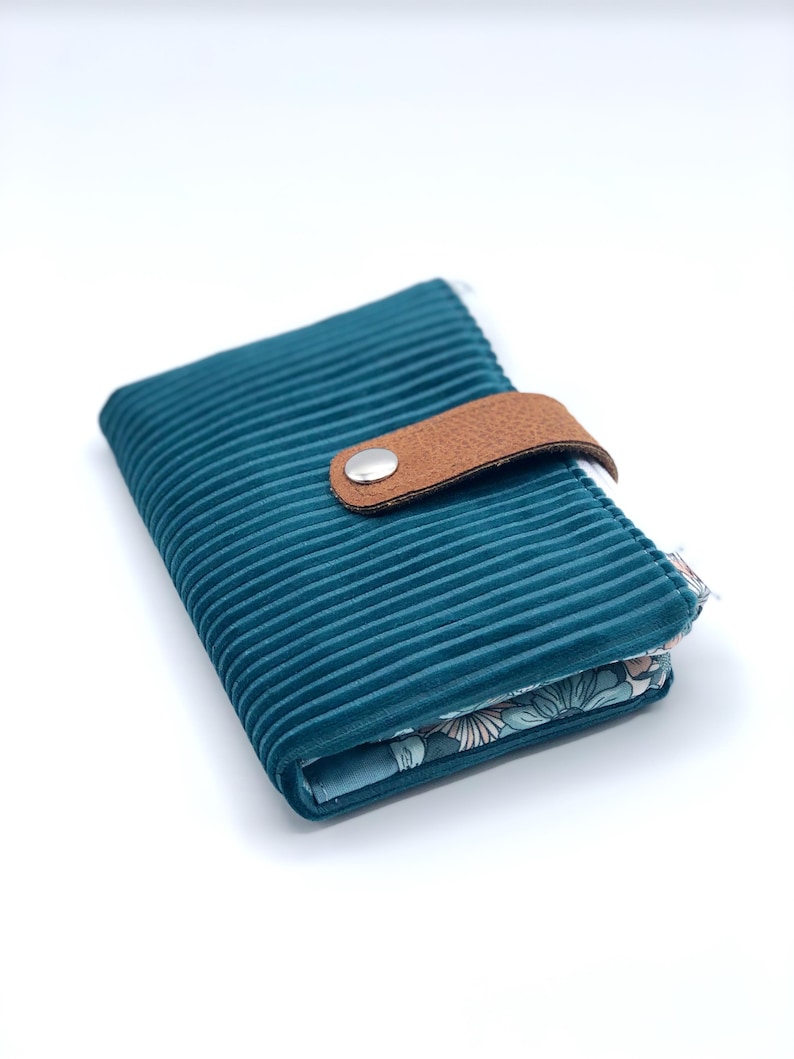 May include: A teal blue corduroy wallet with a brown leather strap and a silver button closure. The inside of the wallet is lined with a floral print fabric.