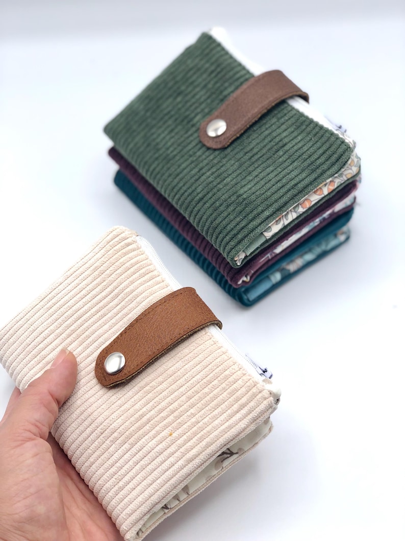 May include: A set of four small wallets made of corduroy fabric. The wallets are in different colors, including green, beige, and blue. Each wallet has a brown leather strap with a metal button closure and a zipper.