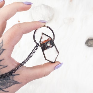 May include: A rose quartz crystal pendant with a copper wire frame and a black chain. The pendant is held in a hand with a floral tattoo.