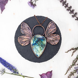 May include: A handcrafted copper pendant necklace featuring a teardrop-shaped labradorite stone with blue and green hues. The pendant has copper wings and a circular bail, displayed on a black slate. The necklace is surrounded by purple flowers and greenery.