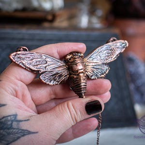 May include: A copper-colored cicada pendant necklace. The insect pendant features detailed wings and body segments. The necklace is held in a hand, showcasing the intricate design and texture of the jewelry. The pendant is attached to a delicate chain.