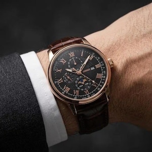 Peut inclure: Une montre-bracelet avec un boîtier couleur or rose, un cadran noir à chiffres romains et un bracelet en cuir marron foncé. Le cadran comporte des sous-cadrans pour la date, le jour et la phase de lune, avec "GRADUE 30 INSATIONS" inscrit.