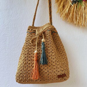 May include: A crocheted tan bucket bag with a drawstring closure and two tassels. The tassels are made of orange and teal yarn and have wooden beads at the top.