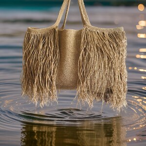 May include: A beige woven tote bag with long fringe detail. The bag has two handles and is suspended above water. The fringe hangs down from the top edges of the bag, creating a textured and stylish look. The bag appears to be made of natural fibers.