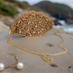 Könnte beinhalten: Eine gehäkelte Halskette in Beige mit einem wellenförmigen Design, verziert mit kleinen weißen Perlen. Die Halskette hat eine zarte goldene Kette. Das Schmuckstück wird auf einer Steinoberfläche präsentiert, mit einem verschwommenen Ozean und Hügel im Hintergrund.