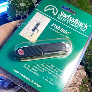 Könnte beinhalten: Ein SwissBuck Fish Tale Taschenmesser in der Originalverpackung. Die grüne Verpackung zeigt die Funktionen des Messers, darunter eine große Klinge, eine kleine Klinge und einen Schraubendreher. Das Messer ist schwarz mit einem roten "BUCK"-Logo.