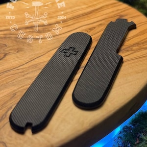 以下が含まれることがあります： 黒いカーボンファイバーテクスチャのポケットナイフ用交換用スケール2つ。1つは十字のシンボル付き。スケールは、木製の表面に「FORGE&BONE CUSTOM」の文字と「2024」の年号が入った円形のロゴとともに表示されています。