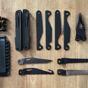 Può includere: Un set di multiutensili neri con vari accessori, tra cui pinze, lame e seghe, disposti su una superficie di legno. È inclusa anche una guaina in nylon nero con il logo "LEATHERMAN". Il set sembra nuovo e inutilizzato.