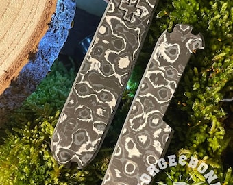 Könnte beinhalten: Zwei schwarze und weiße gemusterte Metallscheiben für ein Taschenmesser. Die Scheiben haben ein Kreuzmuster und eine abgerundete Form. Die Scheiben werden von Forge & Bone Custom hergestellt.