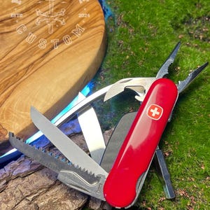 Può includere: Un coltello svizzero rosso con diversi strumenti, tra cui una sega, una lima e un cacciavite, è mostrato aperto. Il coltello presenta una croce bianca su sfondo rosso. Lo sfondo include un tagliere di legno con la scritta "FORGEBONE CUSTOM ESTD 2024".