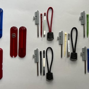 Op de afbeelding: Een set van tien veelkleurige zakgereedschappen met een Swiss Army-logo. De gereedschappen omvatten een rood, blauw en zwart zakmes, een groene en blauwe pincet en een set van vijf gereedschappen met een zwart, rood en groen koord.