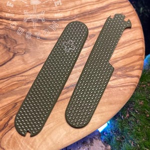 Puede incluir: Dos cachas de cuchillo texturizadas verde oliva con un patrón de diamantes y un logotipo de cruz, sobre una superficie de madera. Una es rectangular, la otra tiene un extremo con muescas. El texto "FORGE & BONE CUSTOM" es visible.