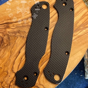 May include: Two black carbon fiber knife scales with a textured weave pattern. The scales have multiple holes for screws and a curved ergonomic design. The text "FORGE & BONE CUSTOM" is visible in a circular logo.