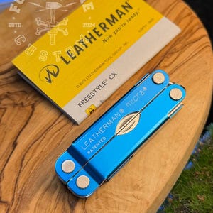 以下が含まれることがあります： 青いLeatherman Micraマルチツールと金色のアクセント、黄色のLeatherman Freestyle CXボックス。マルチツールは開いて木製の表面に置かれています。ボックスには「LEATHERMAN」と「Now you're ready.」と書かれています。
