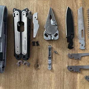 以下が含まれることがあります： 黒いハンドルとグレーのナイロン製の鞘が付いたシルバーのマルチツール。マルチツールが開いており、ナイフ、ペンチ、のこぎり、ドライバーなど、さまざまなツールが表示されています。マルチツールは、黒いナイフ、銀ののこぎり、銀のペンチなど、さまざまな他のツールに囲まれています。