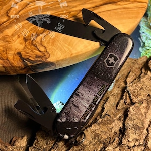 May include: Black multi-tool knife with various implements, including a blade, bottle opener, and screwdriver. The handle features a starry night design with the text "MAN ON THE MOON" and a white cross logo. The knife is displayed on a textured wooden surface.
