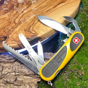 Könnte beinhalten: Ein gelb-schwarzes Multitool-Messer mit mehreren Klingen und Werkzeugen, darunter eine Säge, ein Flaschenöffner und ein Messer. Das Messer trägt den Text "Wenger PATAGONIAN EXPEDITION RACE" und ein rot-weißes Kreuzlogo. Das Messer liegt geöffnet auf einer Holzoberfläche.