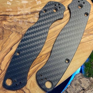 May include: Two carbon fiber knife scales with a woven pattern, featuring multiple holes for screws. The scales are dark gray and have a curved shape, resting on a wooden surface. The text "FORGE&BONE CUSTOM" is visible.