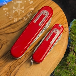 May include: Two red Swiss Army knives, one large and one small, are displayed on a wooden surface. The knives have silver blades and a decorative design on the red casing. The larger knife has a key ring attached.