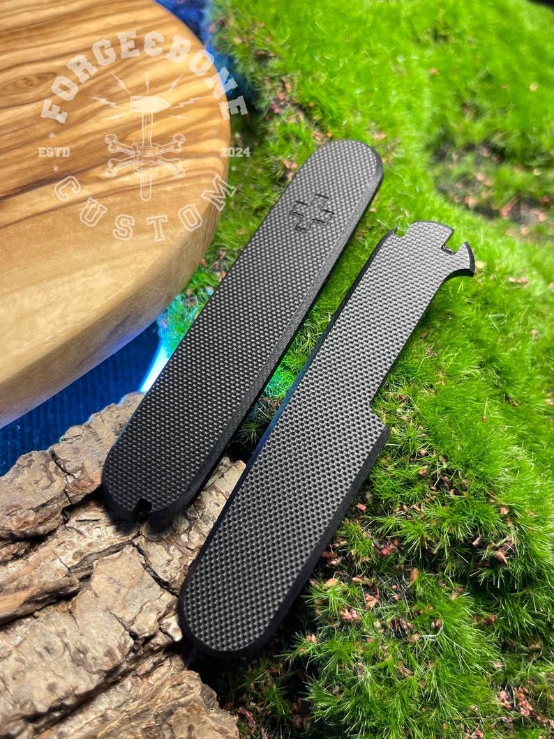 May include: Two black textured custom knife scales with a medical symbol and a notched design. The scales are displayed on a natural wood and moss background. The logo "FORGE BONE CUSTOM ESTD 2024" is visible in the background.