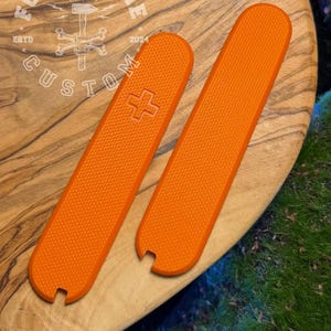 Può includere: Due guance di ricambio arancioni brillanti per un coltello tascabile, con una superficie strutturata. Una guancia presenta un emblema a croce bianca. Le guance sono esposte su una superficie di legno. Il testo "FORGECBONE CUSTOM" è visibile.