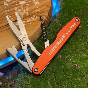 Puede incluir: Una multiherramienta naranja Leatherman Juice CS3 con tijeras, un sacacorchos y otras herramientas. La herramienta está abierta, mostrando las tijeras. La imagen está colocada sobre un fondo de musgo verde y una superficie de madera con el texto "FORGE&BONE CUSTOM ESTD 2024".