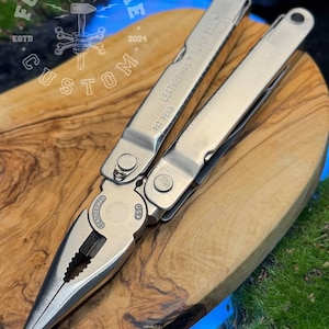 以下が含まれることがあります： 「LEATHERMAN SUPER TOOL」と「USA」の文字が入ったシルバーのマルチツールが、木製の表面に開いた状態で表示されています。このツールには、プライヤー、ナイフ、その他の道具が含まれています。「FORGE&BONE CUSTOM」のロゴが背景に見えます。
