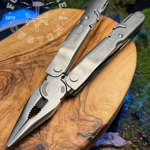 以下が含まれることがあります： ステンレス製のLeatherman Super Tool 200マルチツール。ペンチなどのツールが開いています。「Forge & Bone Custom」の文字と「2024」の年号が入った円形のロゴが刻まれた木製の表面に置かれています。