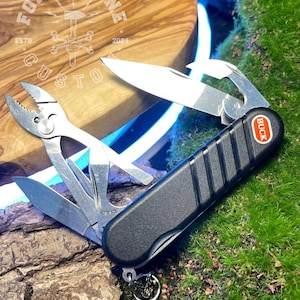 Può includere: Un multiutensile nero con vari strumenti in acciaio inossidabile, tra cui un coltello, pinze e cacciaviti. L'utensile ha un manico nero con il marchio "BUCK" in un cerchio rosso. L'utensile è su muschio verde.