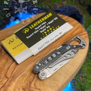 May include: A Leatherman Squirt PS4 multi-tool with a black and silver finish, and a matching product guide. The tool is open, revealing a blade and other implements. The items rest on a wooden surface.