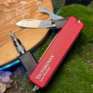 Könnte beinhalten: Ein rotes Victorinox SportRatchet Multi-Tool mit silberner Klinge und schwarzem Schraubendreheraufsatz. Das Werkzeug ist geöffnet und zeigt die Klinge und einen Schraubendreherbit. Der Hintergrund umfasst eine Holzoberfläche und grünes Moos.