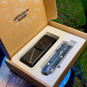 May include: A Leatherman Crunch multi-tool and a black leather sheath housed in a cardboard box. The box lid features the text FORGEBONE, LEATHERMAN GARAGE, and TOM. The multi-tool is silver-coloured metal with the text LEATHERMAN CRUNCH visible.