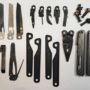 Op de afbeelding: Een assortiment vervangende onderdelen en componenten voor multitools, waaronder messen, vijlen en diverse gereedschappen. De onderdelen zijn voornamelijk zilver en zwart, met verschillende vormen en maten. De afbeelding is een bovenaanzicht van de gereedschappen.